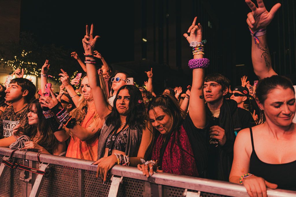Concert crowd raises hands and cheers enthusiastically at a dimly lit music venue during a live performance.