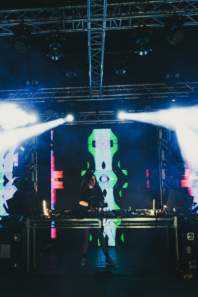 Electronic music performance stage with colorful geometric visuals and bright lighting effects against a dark backdrop.