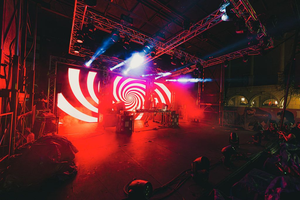 Red neon lights illuminate a dark nightclub stage setup with metal truss rigging and sound equipment.