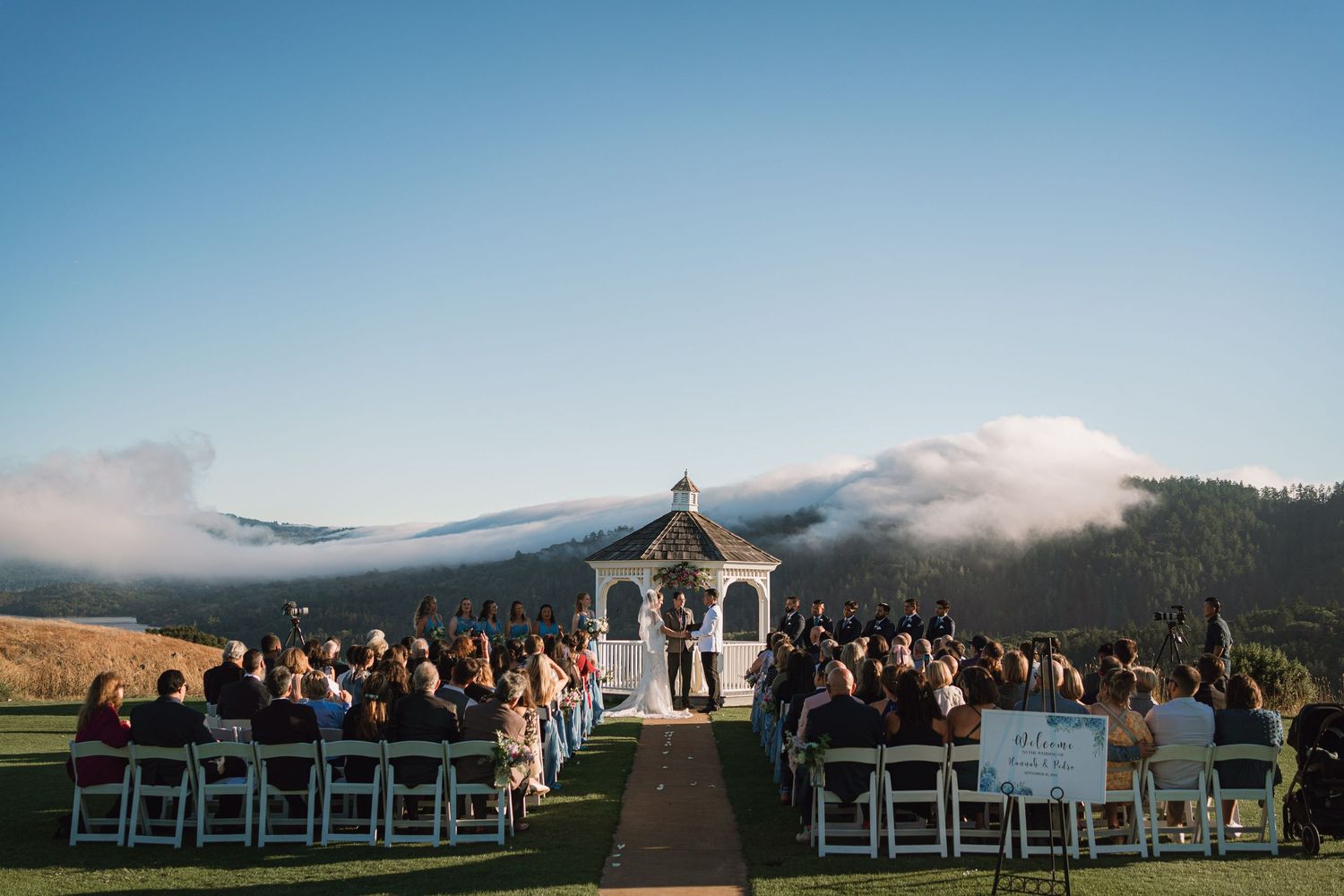 An outdoor wedding ceremony at a gazebo with mountain views and low-lying clouds in the background.