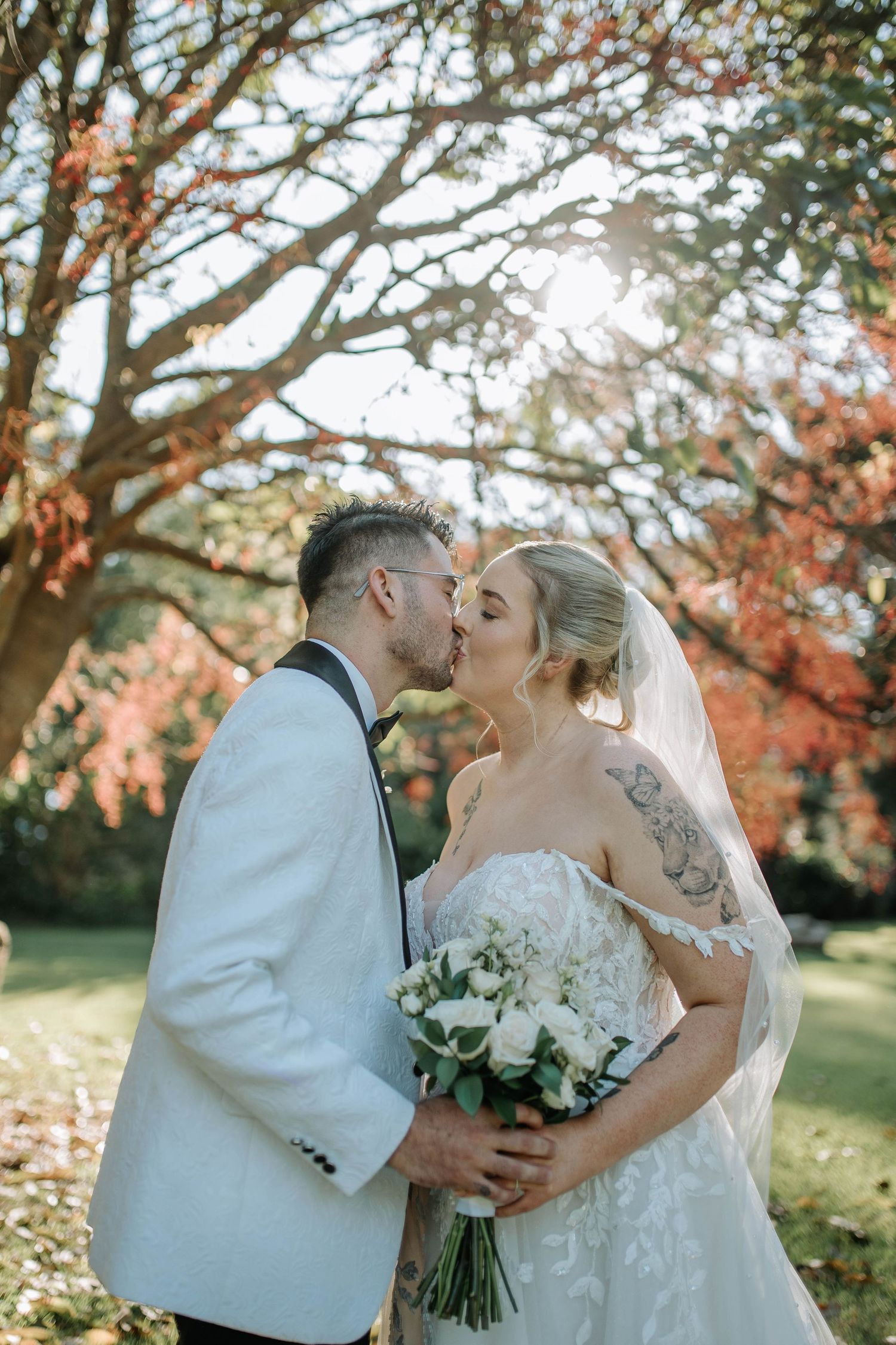 A romantic wedding couple shares a kiss under autumn trees while holding a white floral bouquet.