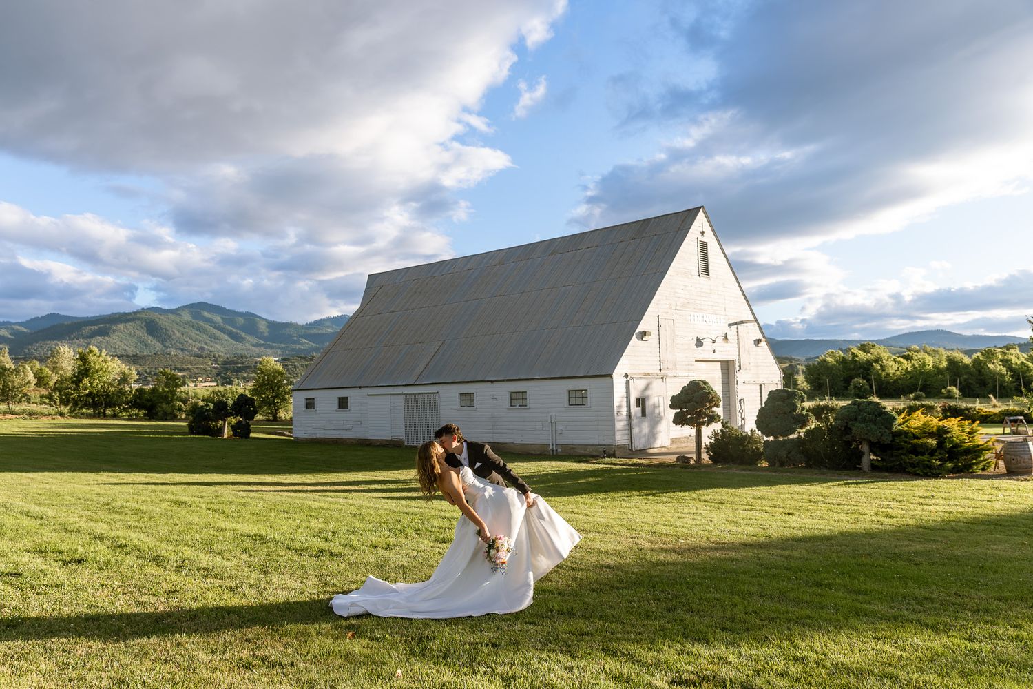 Bride and groom share a romantic dip kiss on lush green lawn before a white barn with mountain backdrop.