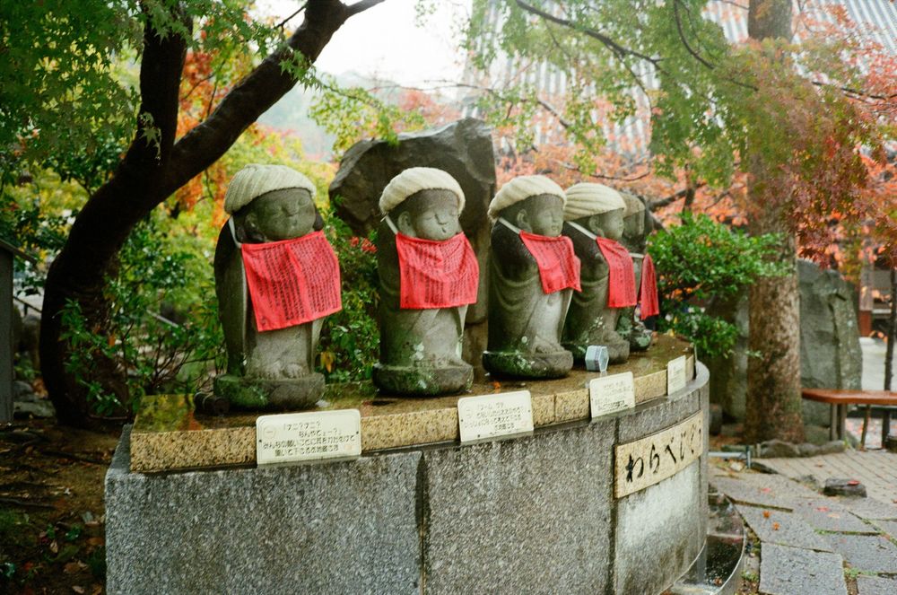 Row of stone statues with red caps along a garden wall surrounded by lush green trees and foliage.