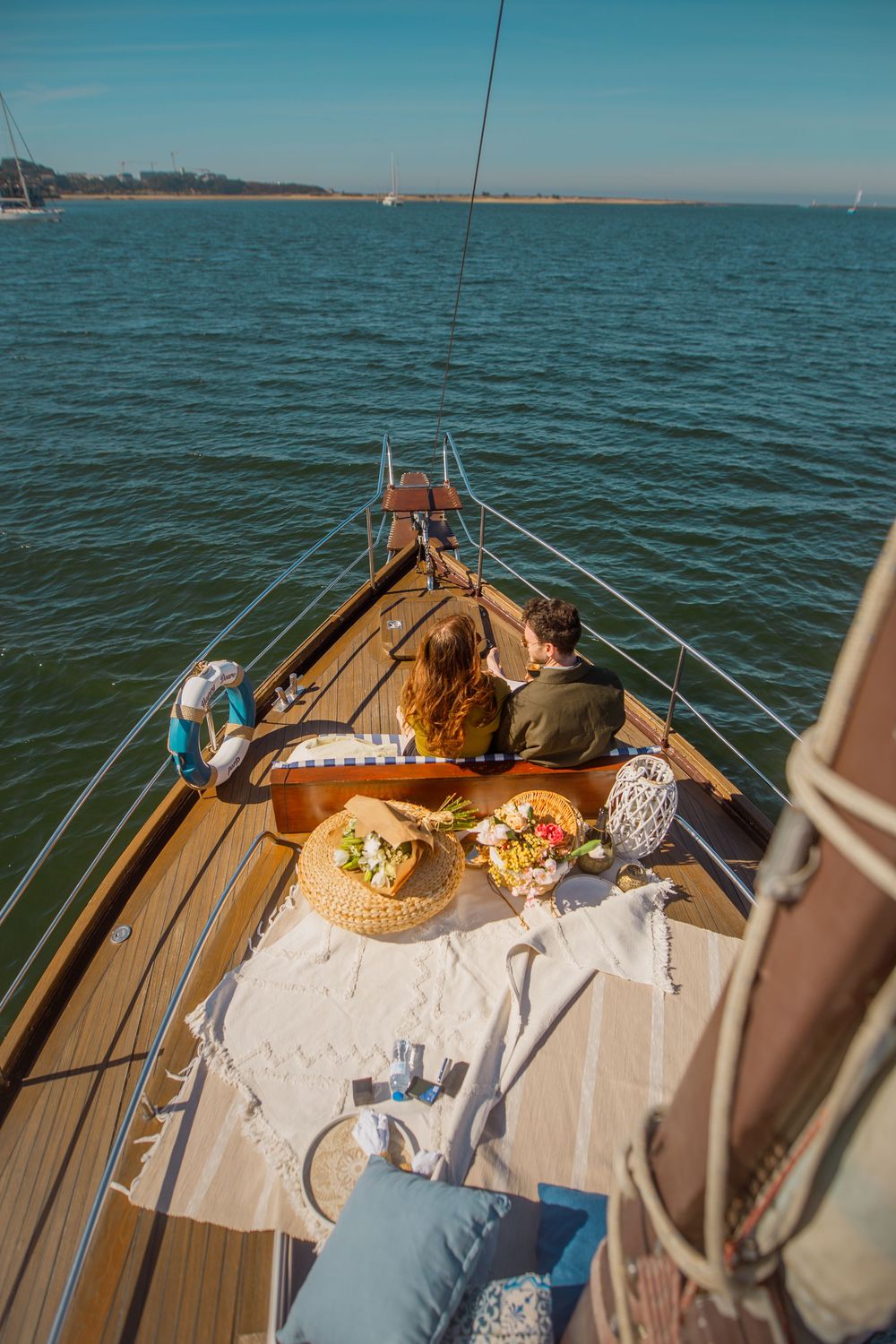A romantic picnic setup on the bow of a wooden sailboat with food and cushions while cruising on turquoise waters.