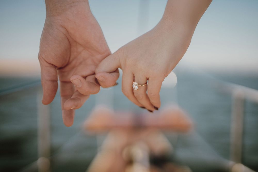 Close-up of hands wearing wedding rings against a blurred ocean background in soft natural light.