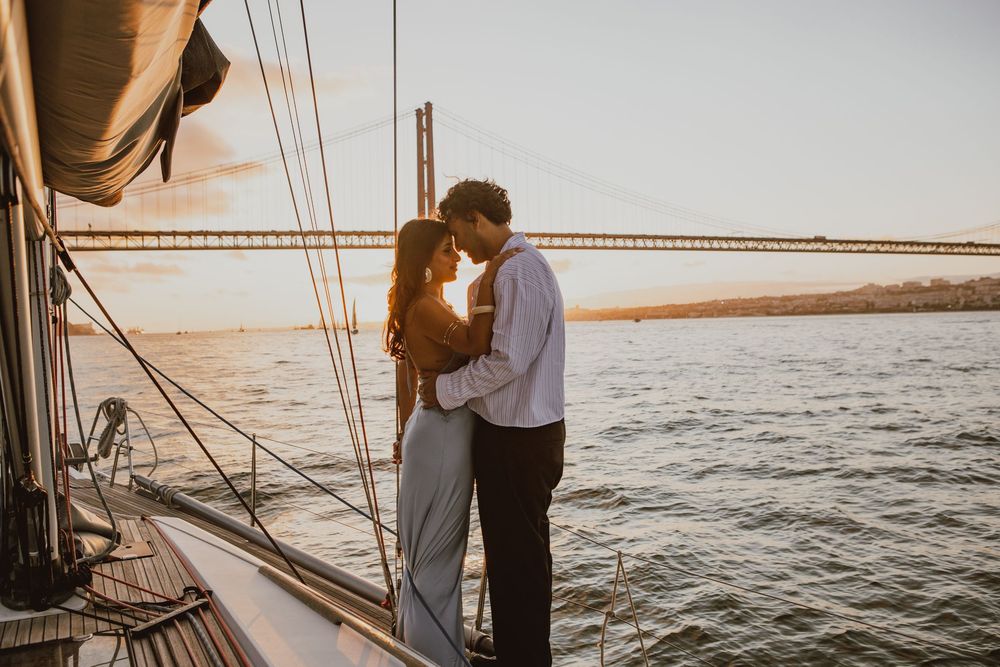 A couple embraces on a sailboat deck during golden hour with water and coastline in the background.