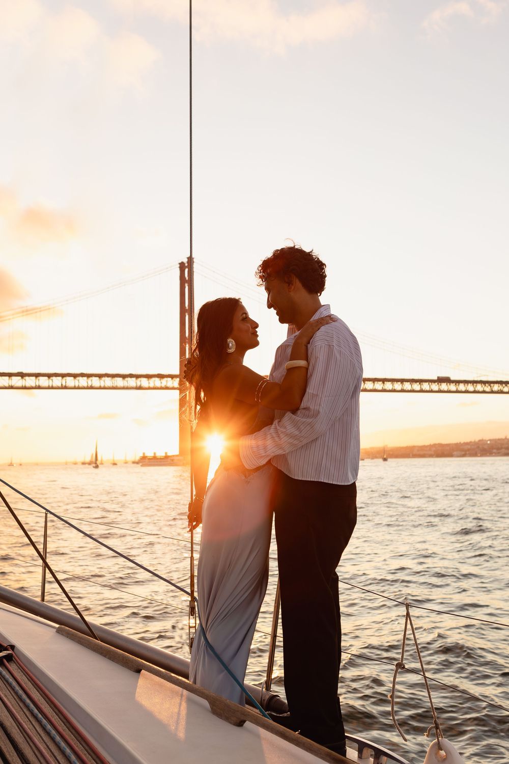 Romantic couple embracing on a boat at sunset with a bridge visible in the background over calm waters.