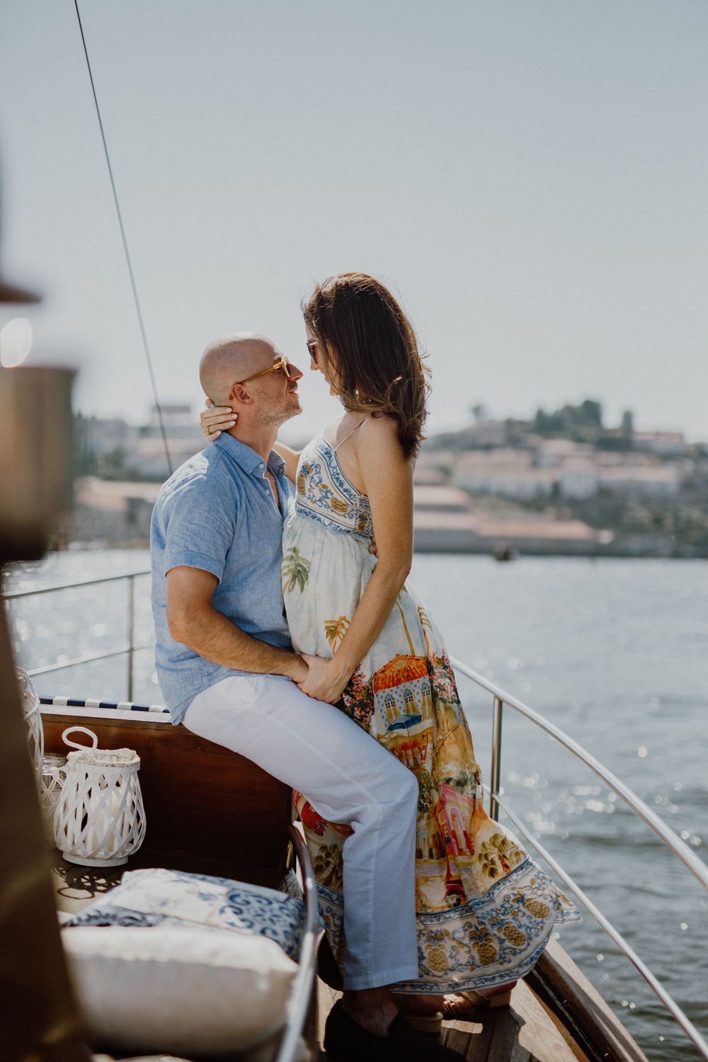 A romantic couple embraces on a sailboat during sunset with a waterfront cityscape in the background.