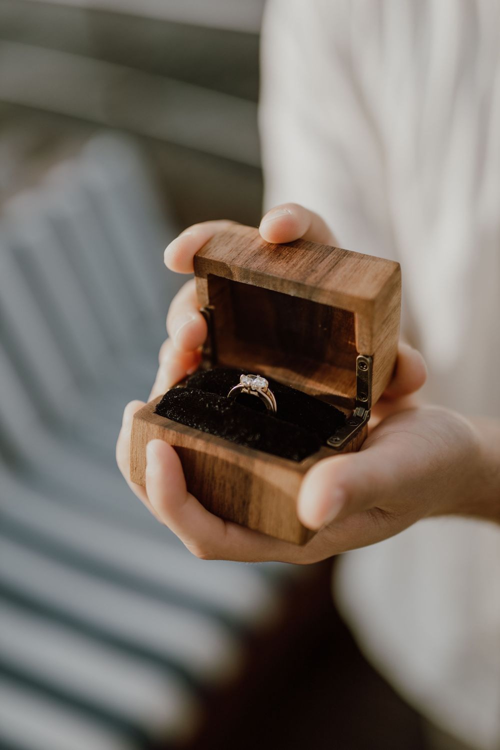 Hands holding a small wooden box with an engagement ring inside, photographed in soft natural light.
