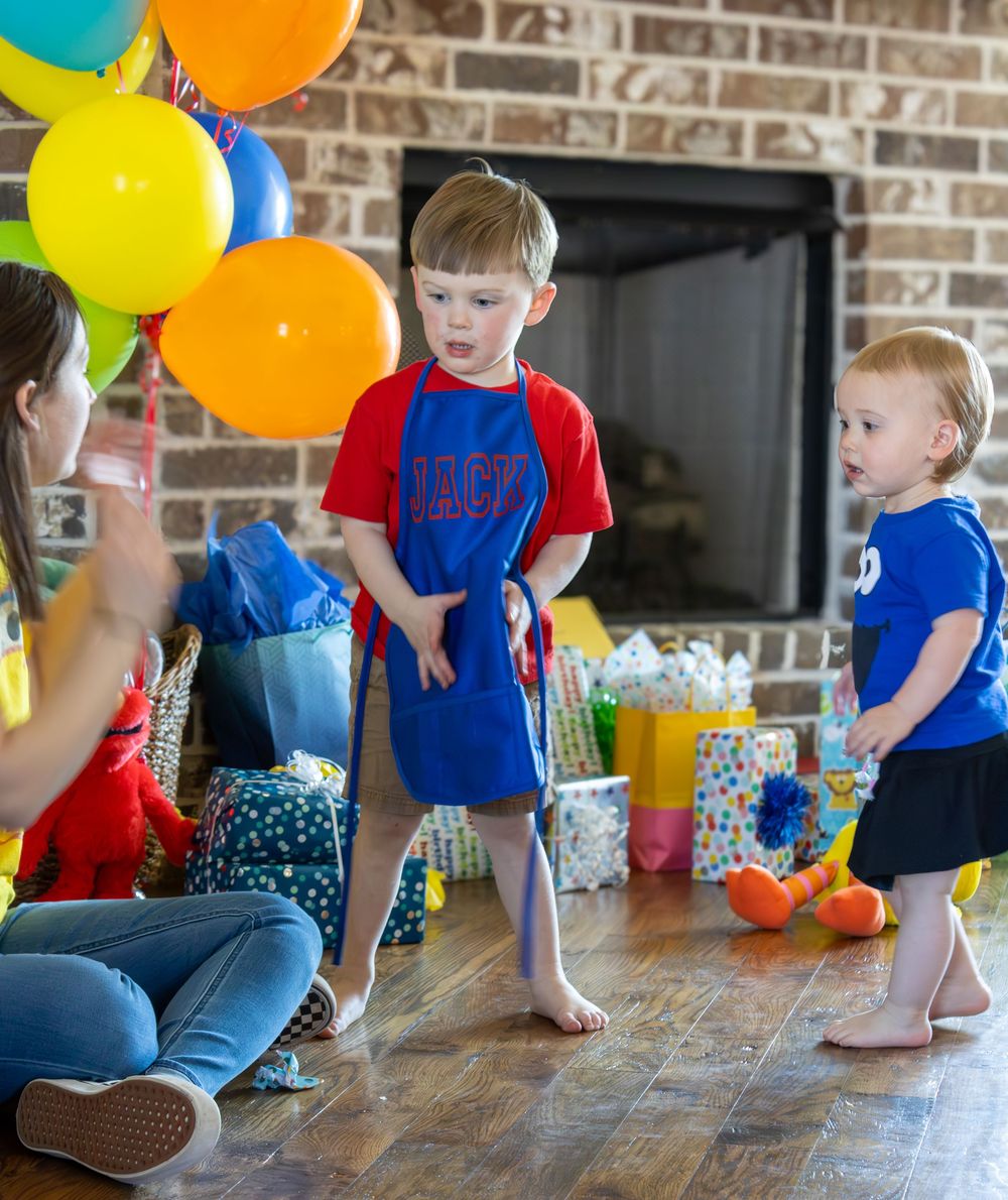Children in blue and red shirts play together with colorful balloons on a hardwood floor near a brick wall.