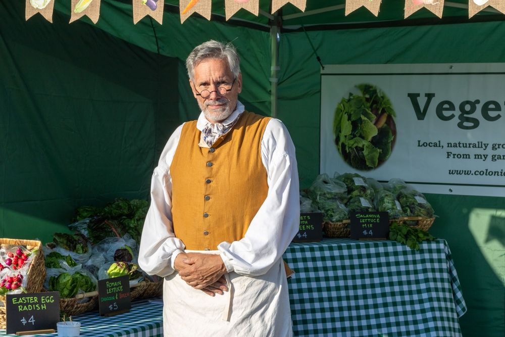 A person in traditional Bavarian attire serves food at an outdoor market stand with checkered tablecloths and signage.