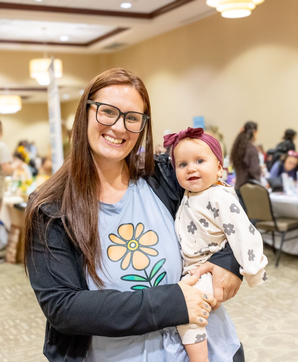 A caregiver holds and smiles at a baby wearing a floral headband in a community gathering space.