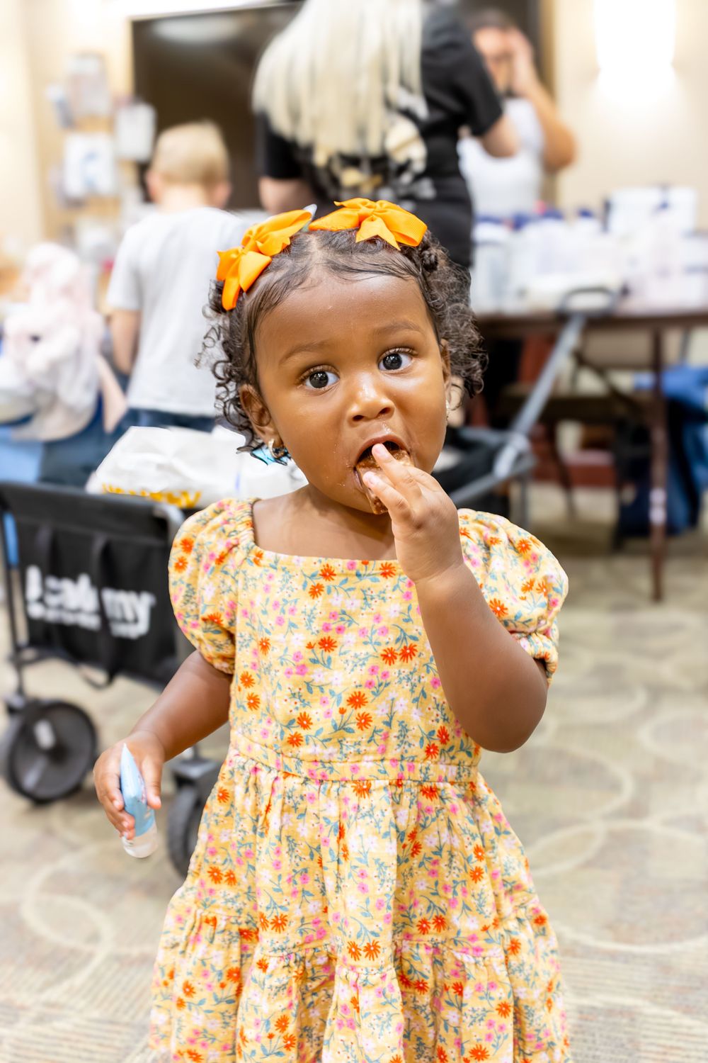 A child in a floral dress with an orange bow explores their surroundings at an indoor event.