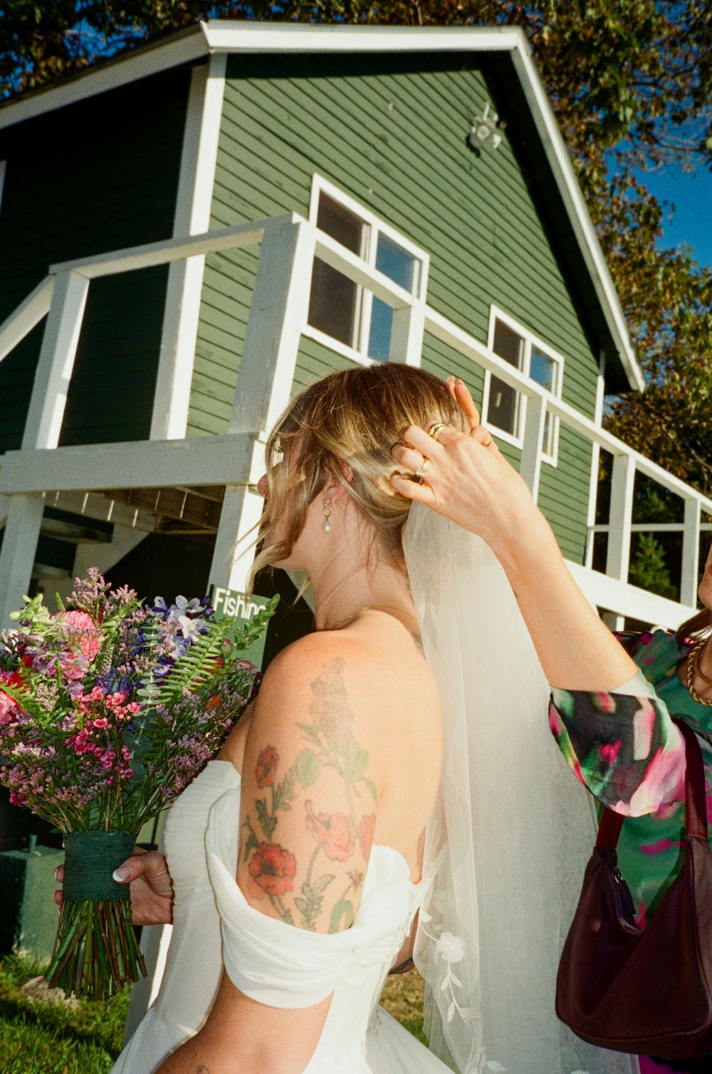 Bridesmaid assists in repositioning the bride's veil while the bride holds her floral bouquet outside before their wedding ceremony in Maine