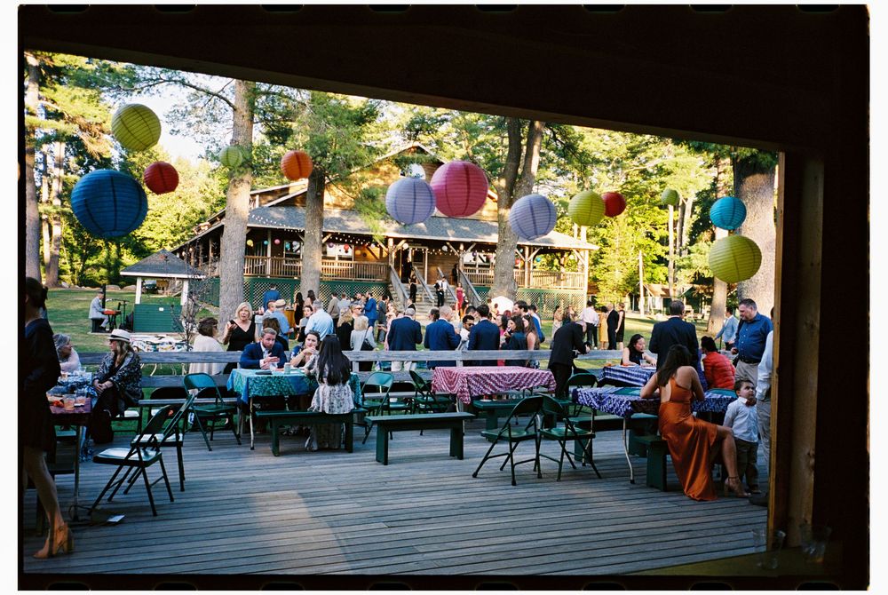 Scene of all the wedding guests celebrating after a late summer ceremony underneath colored paper lanterns at Kamp Kohut in Oxford, Maine.