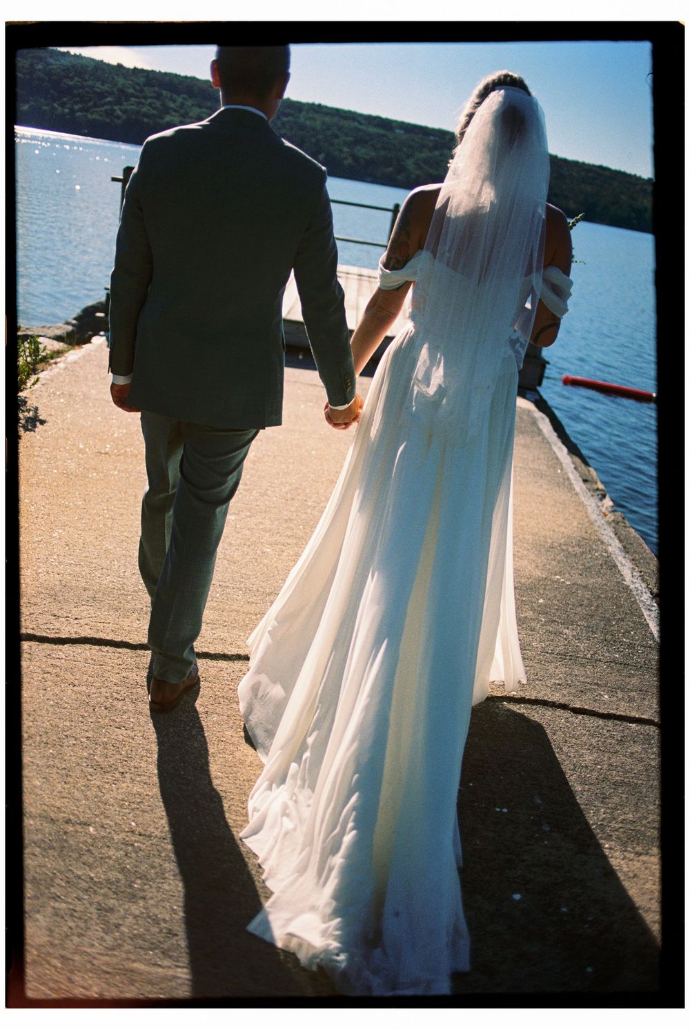 Couple holds hands walking together away from the camera down a dock overlooking a large lake before their summer wedding ceremony in Maine.