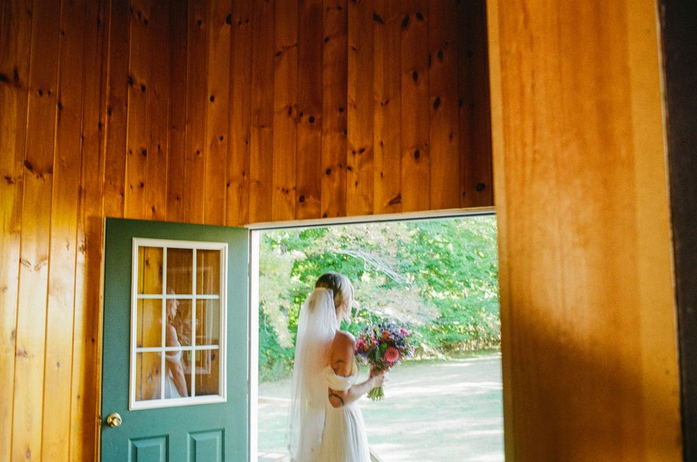 A quiet moment showing the bride and her floral bouquet looking through a door into the grass before her wedding ceremony in Oxford, Maine.