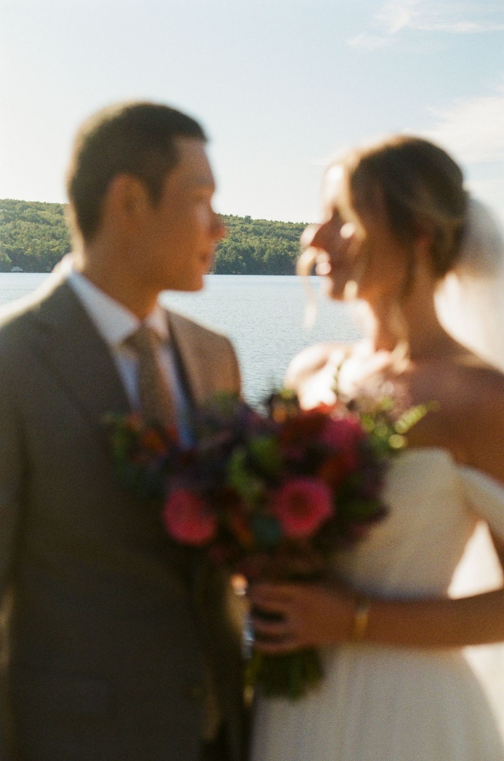 A portrait of the wedding couple in front of a lake with the background in focus and the lovebirds blurred but looking lovingly at eachother