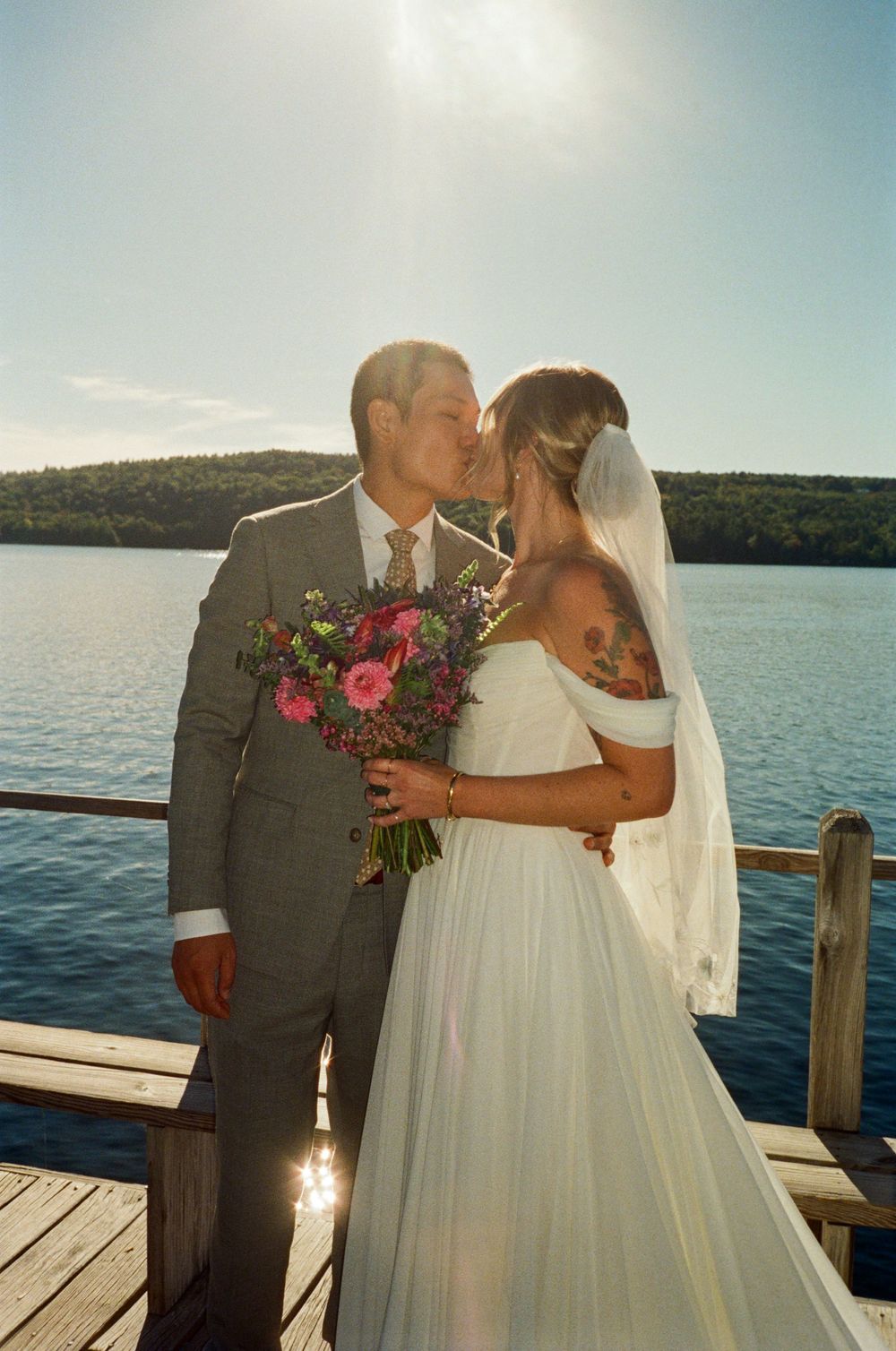 Couple kisses in the sunshine at the end of a wooden dock by a lake before their wedding ceremony in the summer in Oxford, Maine.