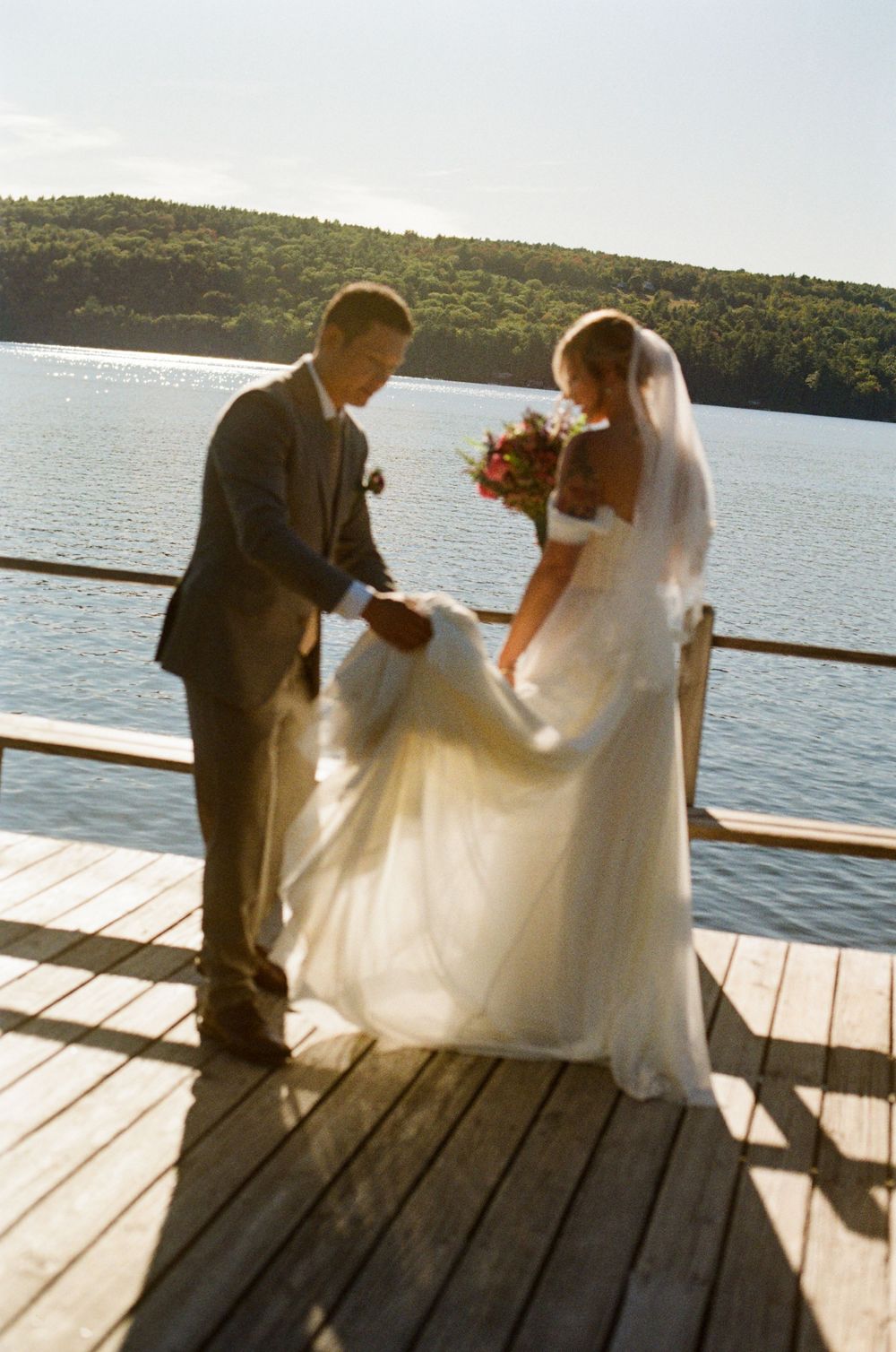 The groom helps untangle the brides's dress on a dock by a lake as the sun shines through the fabric. A quiet moment between bride and groom