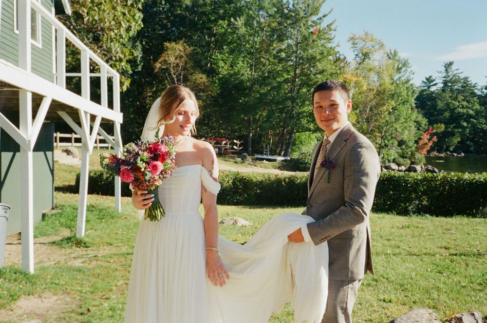 The bride looks at the groom while he holds the train of her dress and smiles at the camera. Candid portrait of the couple before ceremony.