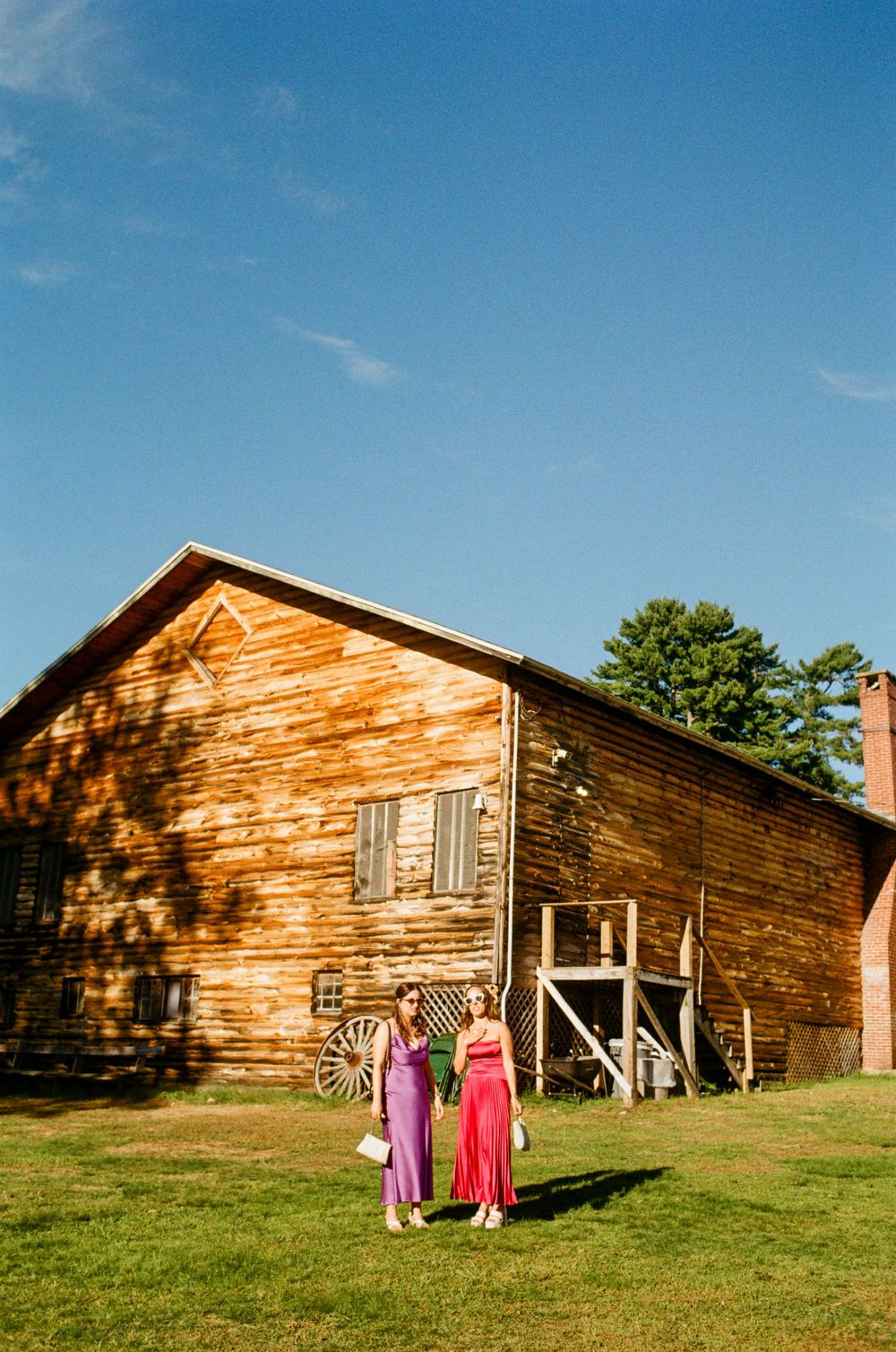 Two bridesmaids stand in colorful dress in the sunshine in front of a large wooden barn before their friend's wedding ceremony on 35mm film.