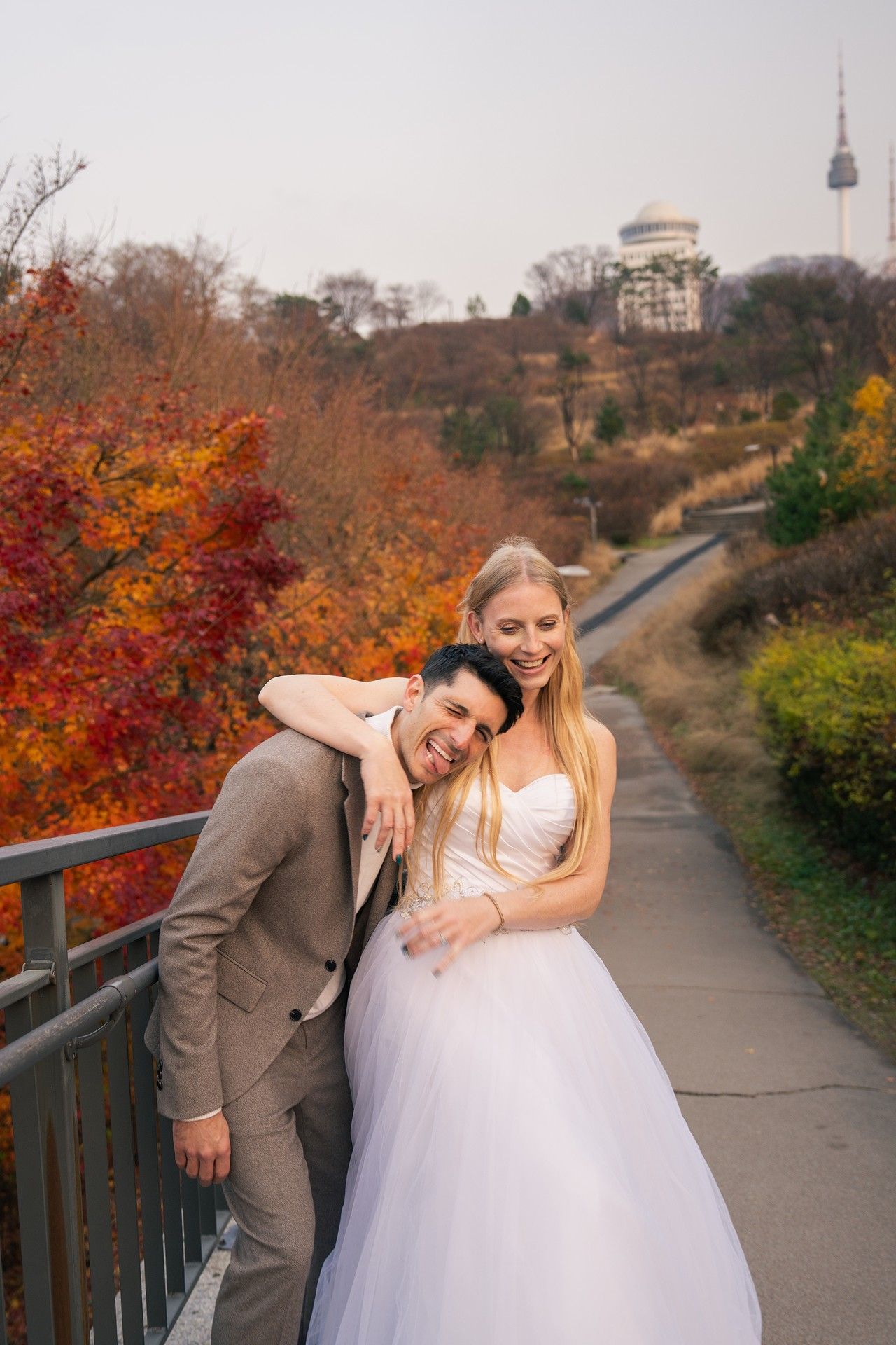 A couple in wedding attire poses playfully on a path surrounded by vibrant autumn foliage and trees.