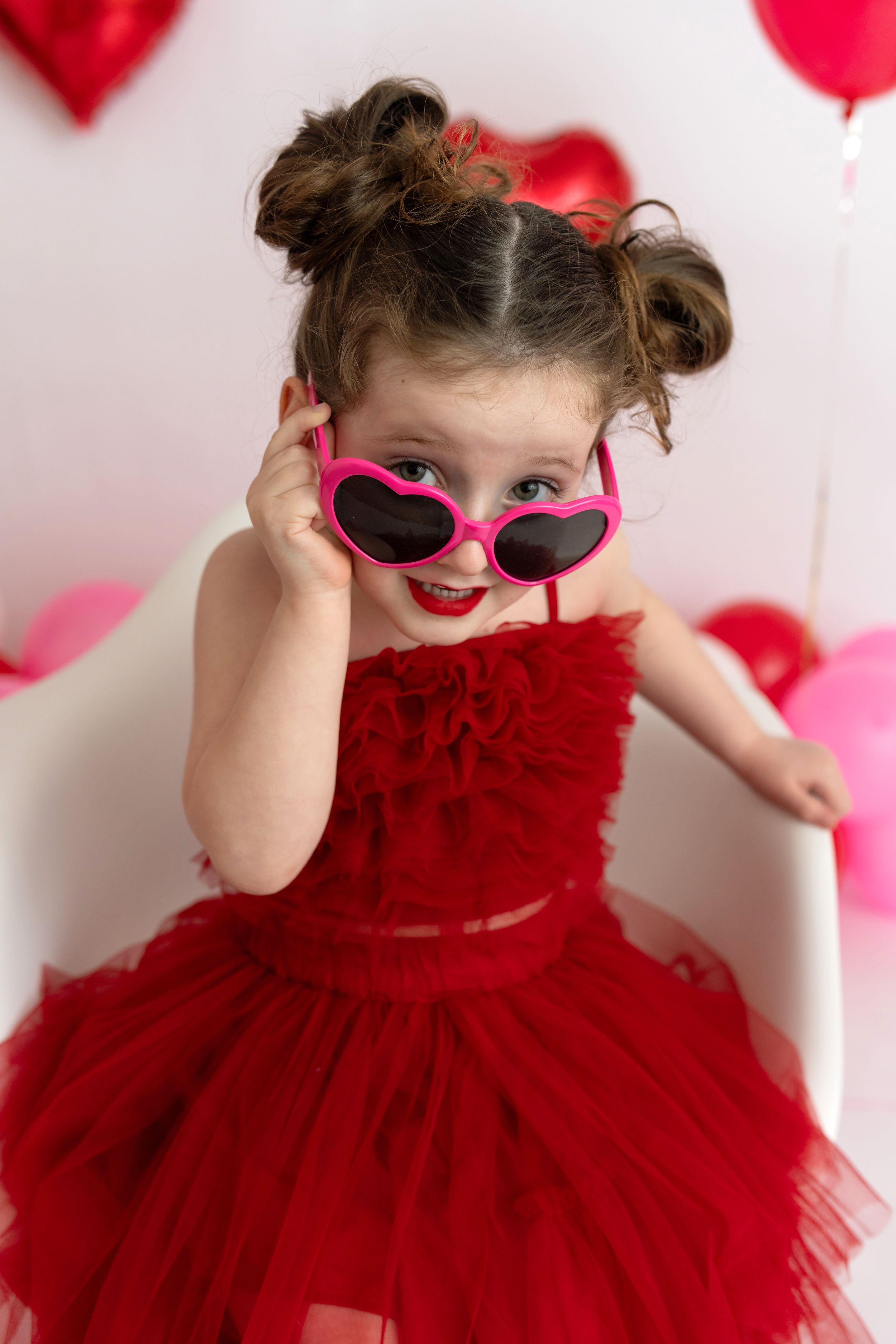 Little girl in a red dress sitting in a white chair pulling down her red heart sunglasses