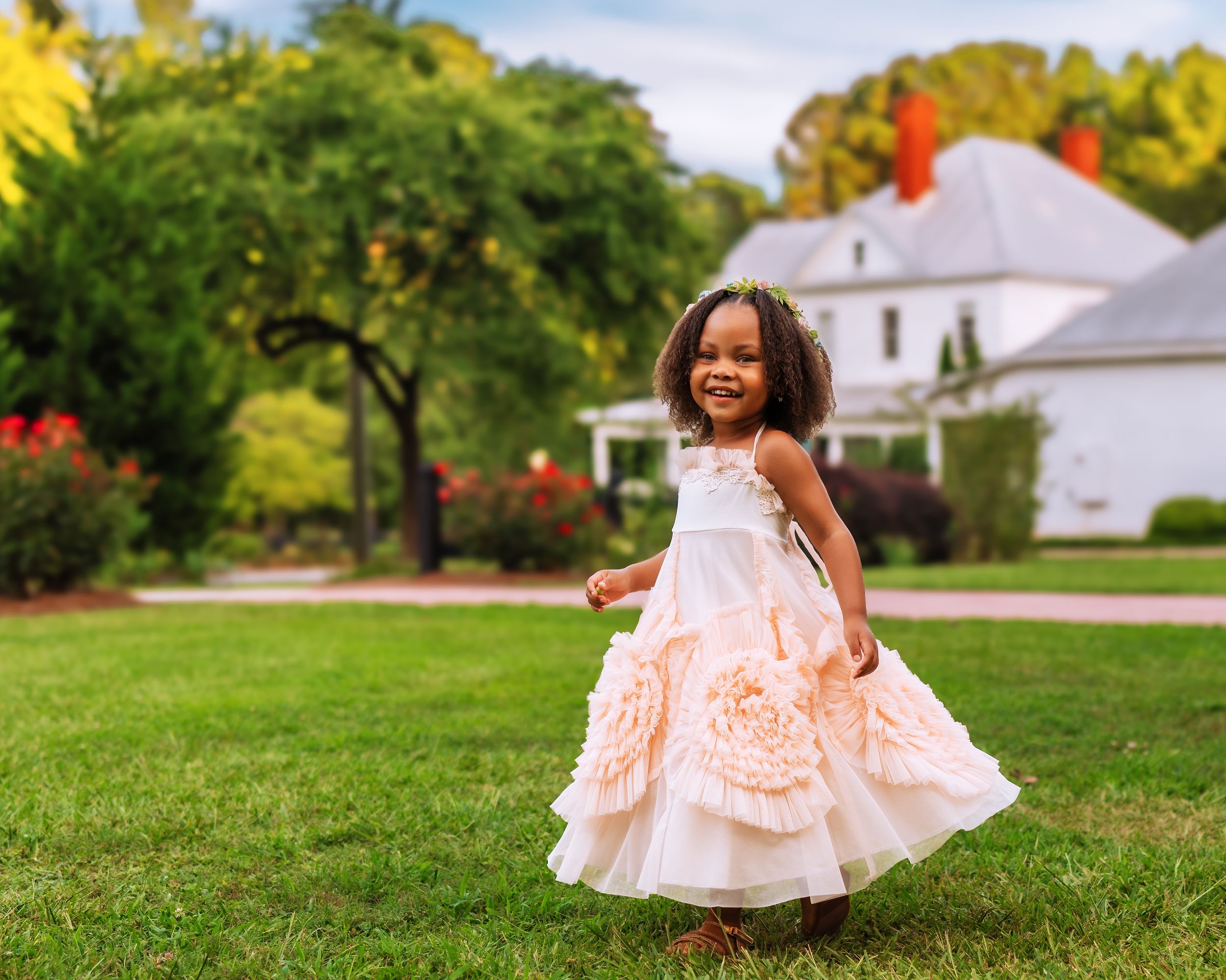 Little girl in a peach dress standing on a green lawn in front of a white historic house call the mcgarity house.