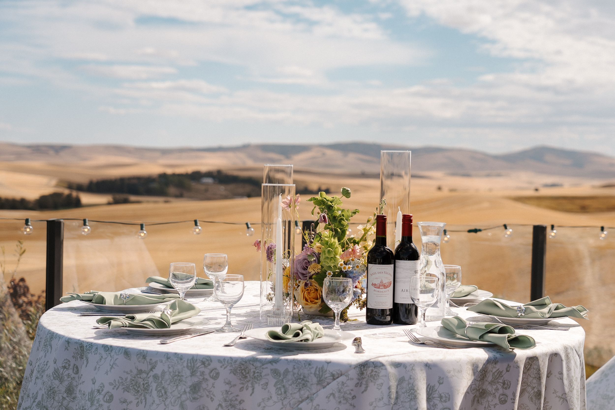 Elegant outdoor dining tables set against rolling hills with wine glasses and place settings at sunset.