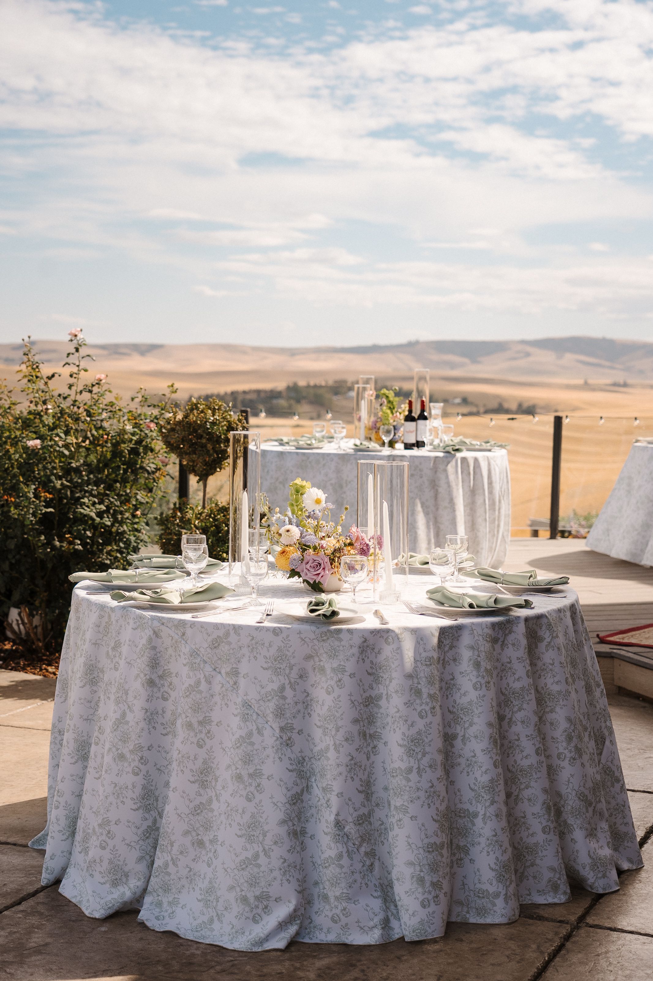 Elegant round table with white damask tablecloth set for dining outdoors overlooking rolling hills and desert landscape.