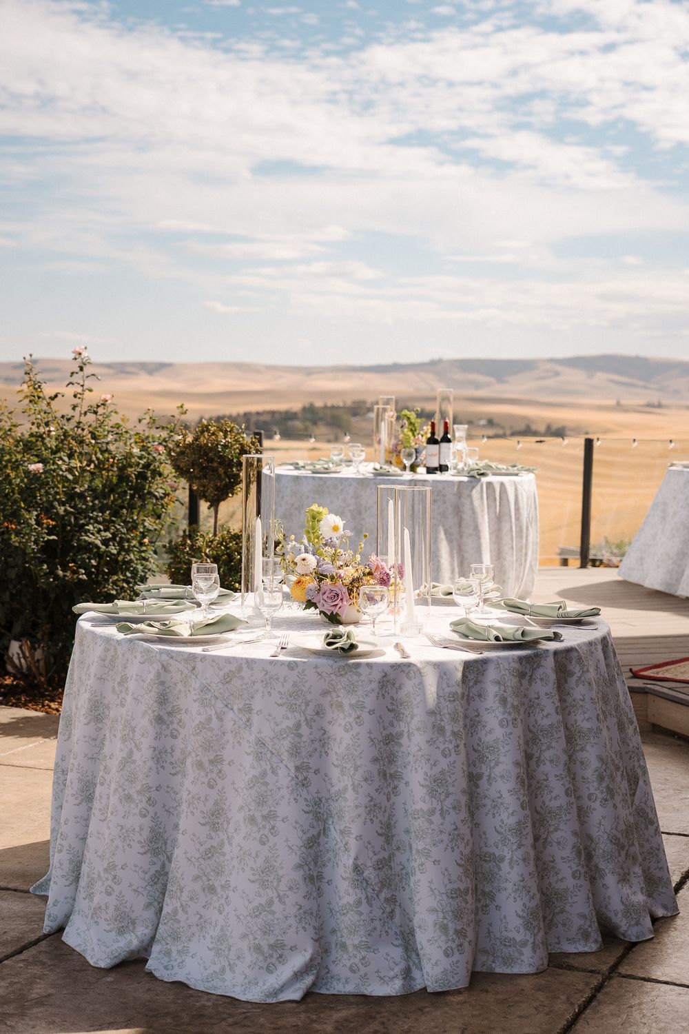 Elegant round table with white damask tablecloth set for dining outdoors overlooking rolling hills and desert landscape.