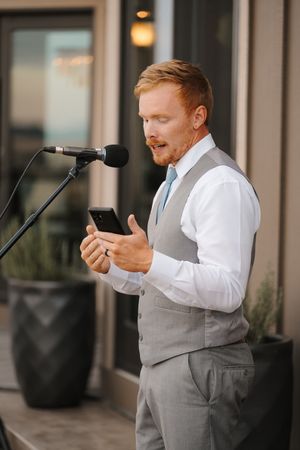 A person in a white dress shirt and gray vest reads from their phone during what appears to be a wedding ceremony.