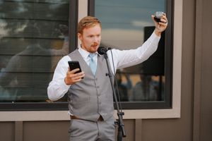A person in a grey vest and white shirt holds up two phones while standing outside a building.