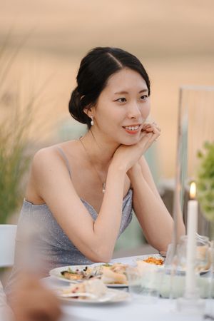 A person in a grey dress smiles while dining at an elegant table setting with soft lighting and blurred background.
