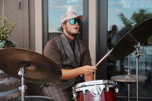 Drummer wearing a baseball cap performs on a red drum kit in front of a glass window.