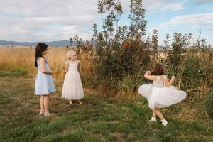 People in white dresses walking through a scenic field with tall grass and trees at golden hour.