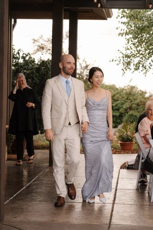 Couple walking arm in arm through a covered patio area wearing light colored formal attire.