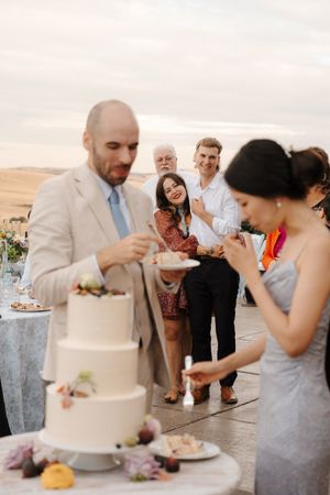 Wedding guests gather around an elegant white wedding cake at an outdoor reception.