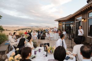 Wedding reception guests seated at outdoor tables on a terrace overlooking desert landscape at sunset.