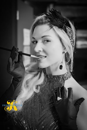 Black and white portrait of a glamorous 1920s style figure in a beaded dress and long gloves holding a cigarette holder.