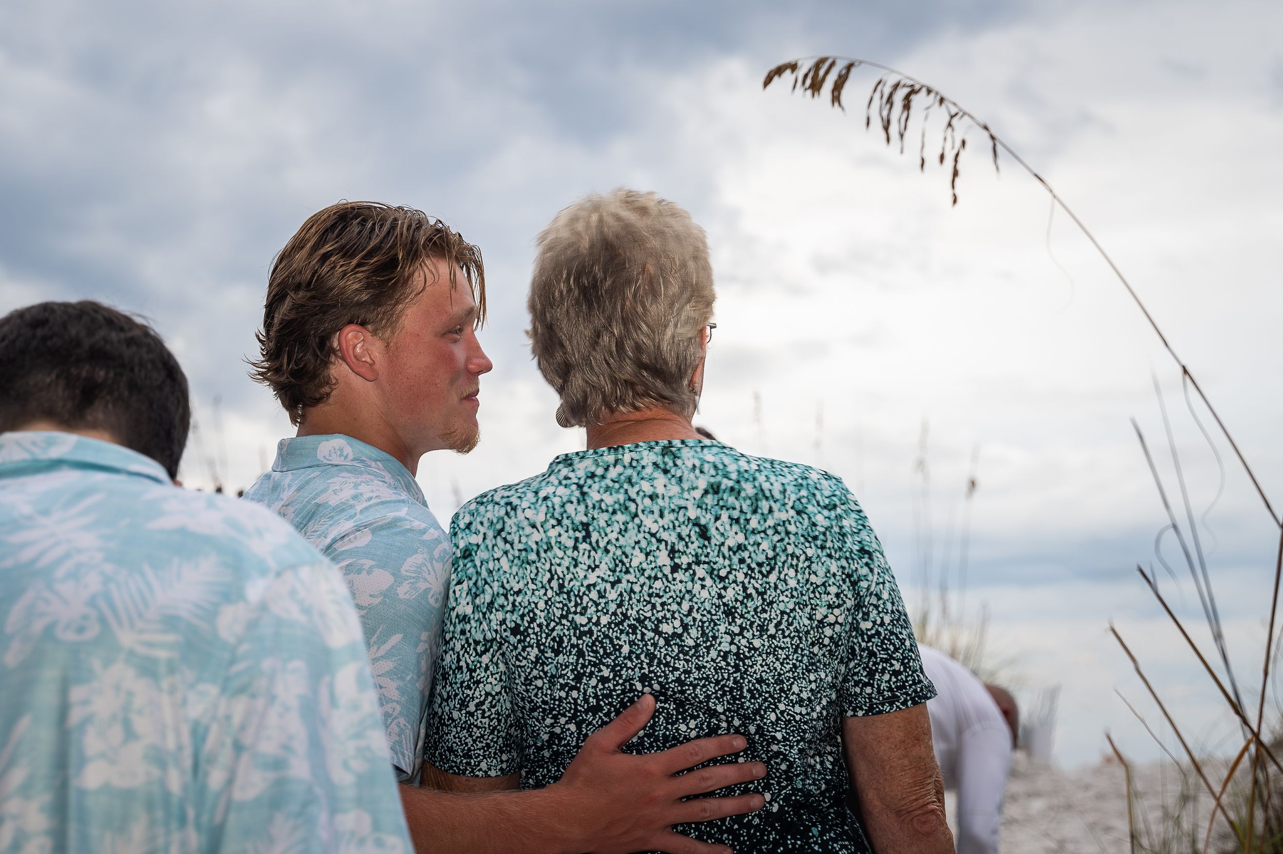 A couple embraces against a cloudy sky backdrop with tall grass in the foreground.