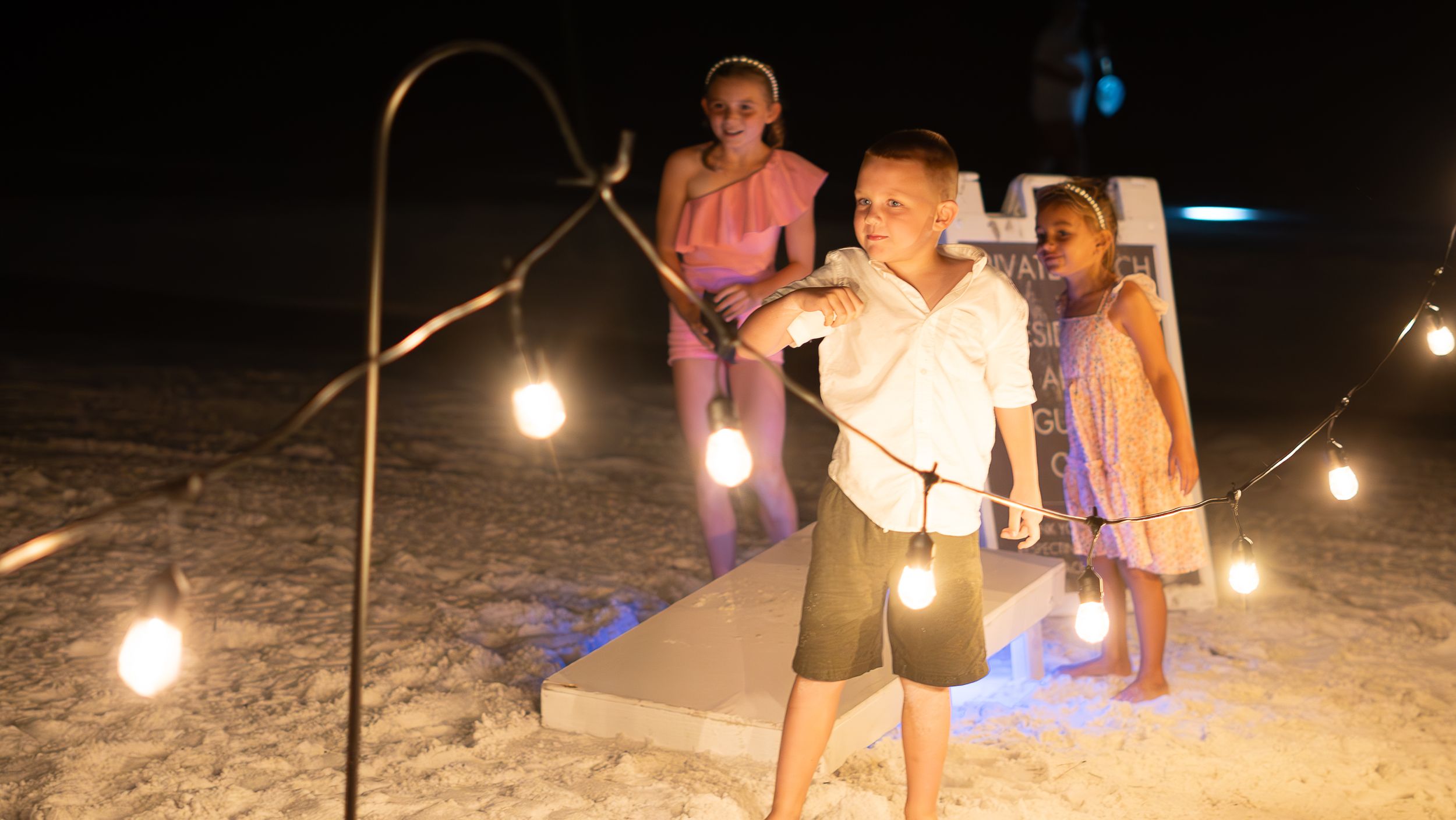 Illuminated string lights create a warm glow over a beach wedding ceremony setup at dusk.