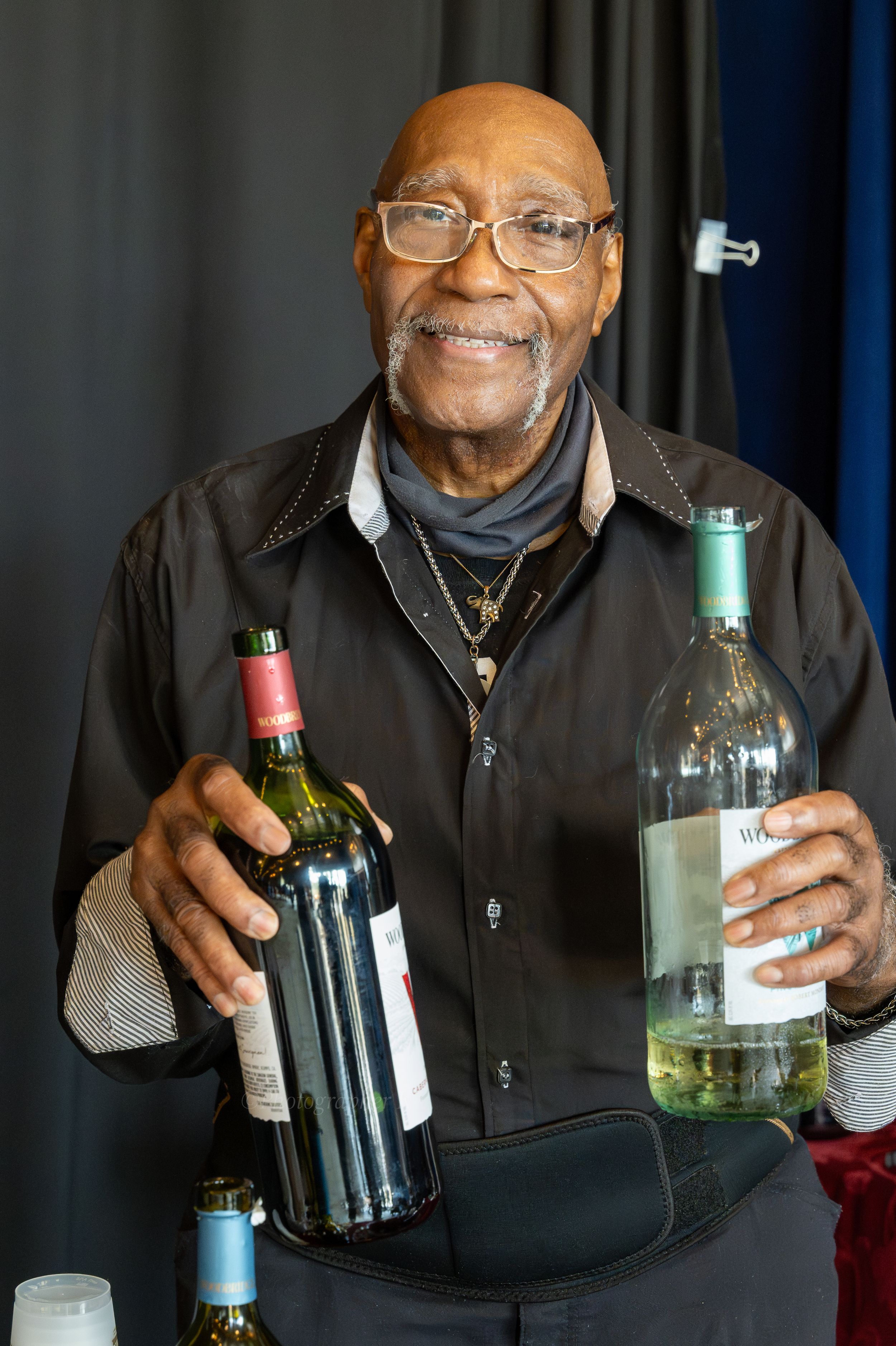 A smiling senior wearing glasses and a black jacket holds up various wine bottles in a sequence of photos.