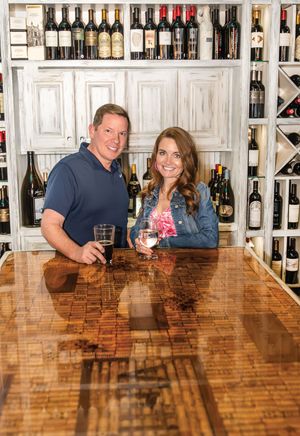 A couple sits at a long wooden bar counter with wine bottles displayed on shelves behind them in a rustic tasting room.