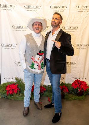 Two people pose at a Christie's event with poinsettias and a holiday backdrop, one wearing a cowboy hat and festive sweater.