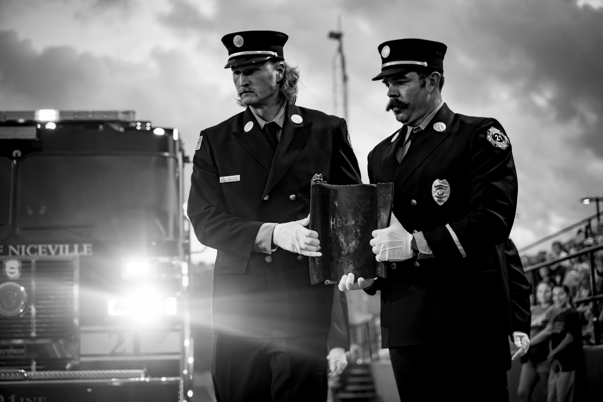 Two firefighters in dress uniforms carry equipment near a Niceville fire truck in a dramatic black and white photo.