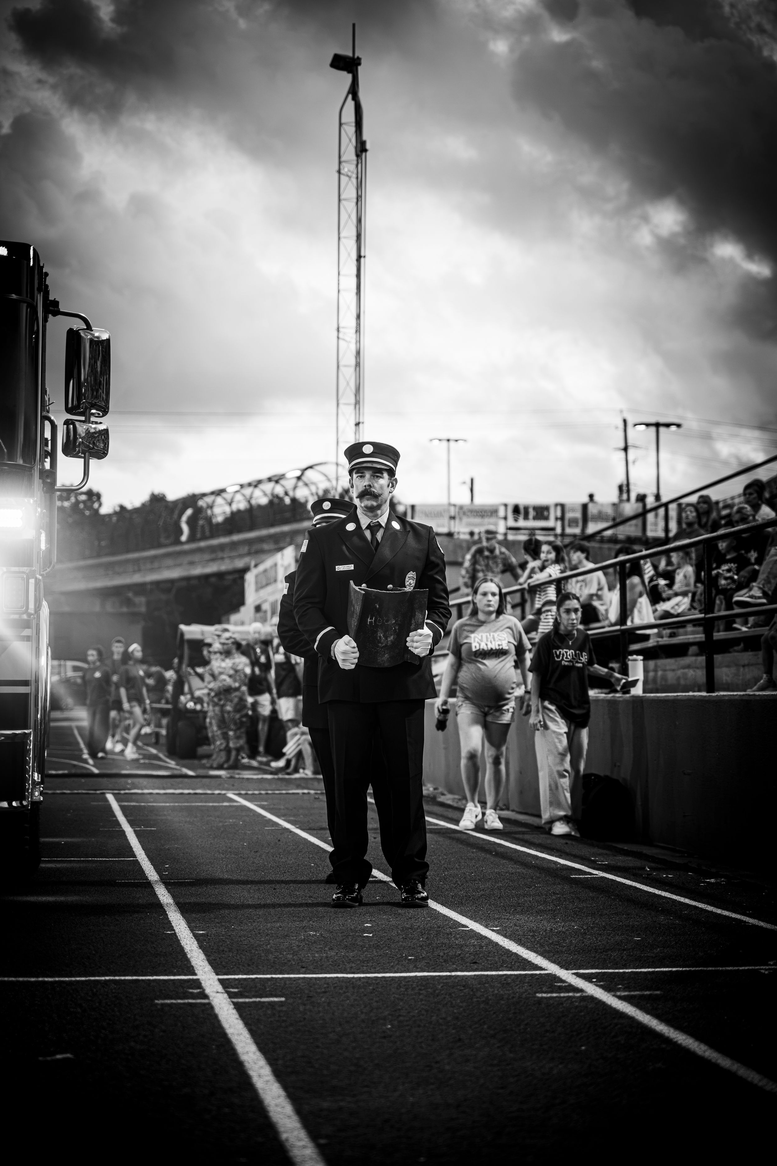 Black and white photo of a uniformed firefighter marching on a track at a stadium event under dramatic cloudy skies.
