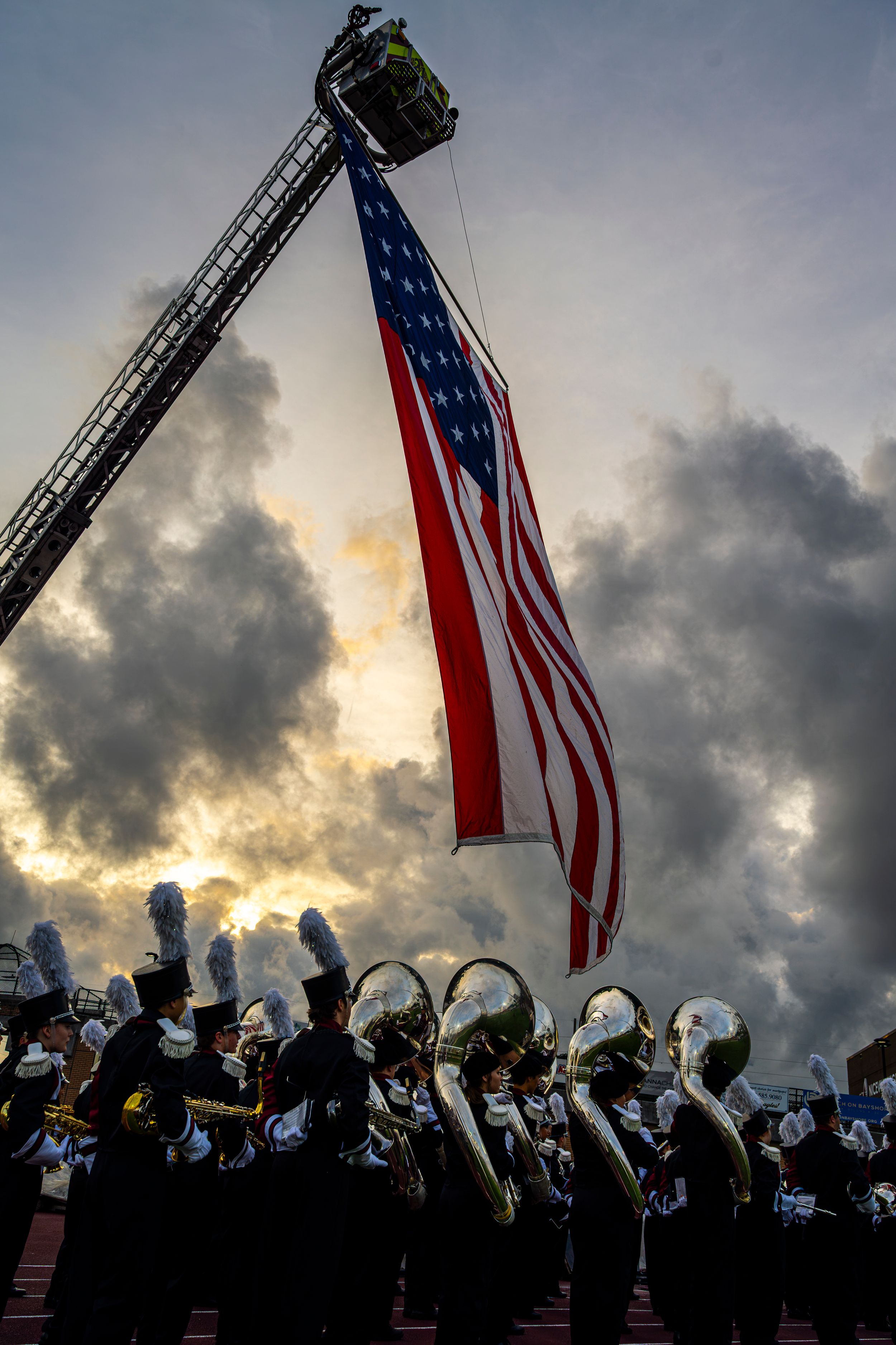 Marching band performs under a large American flag hung from a fire truck ladder against a dramatic cloudy sunset sky.
