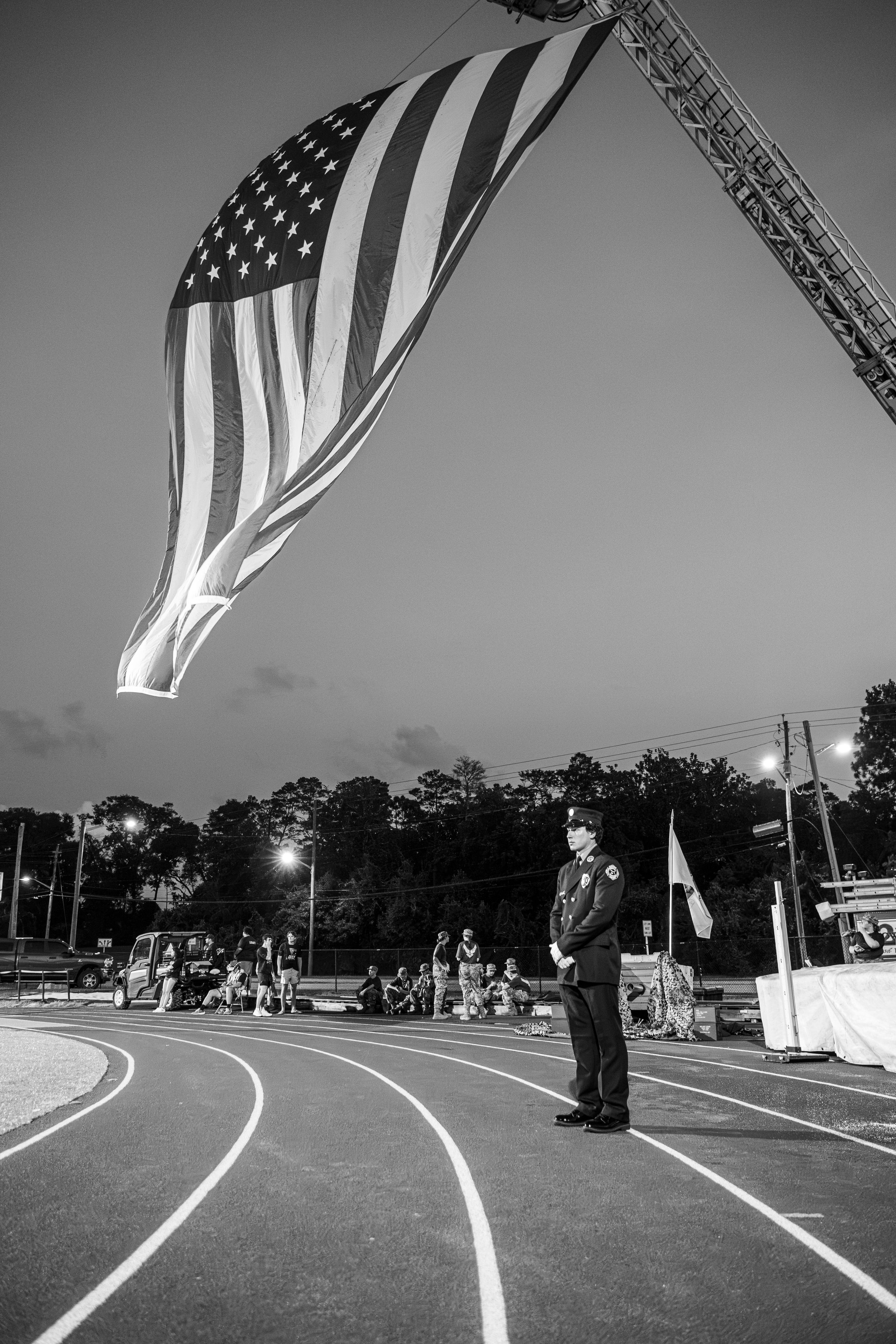 Black and white photo of officer standing at attention under American flag on a track at dusk.